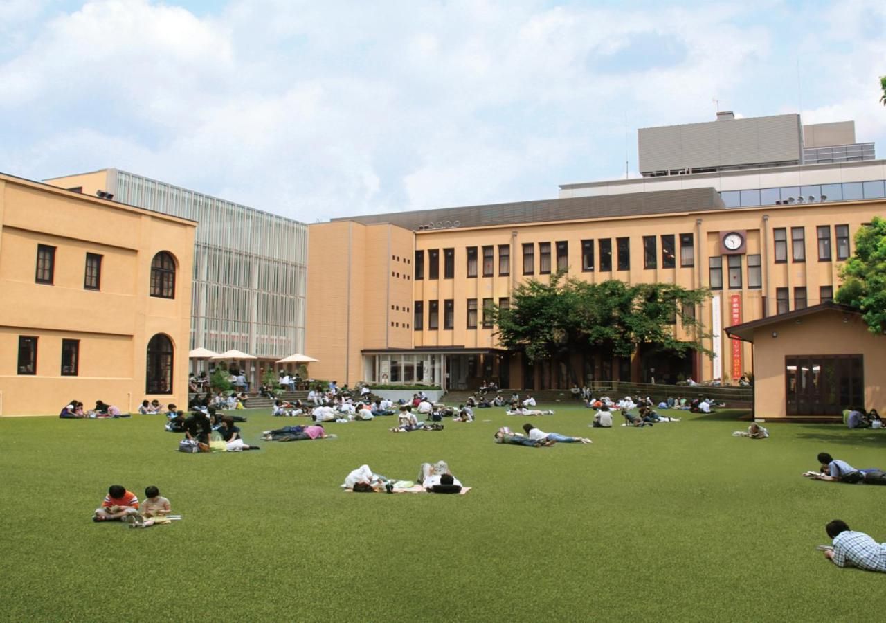 A courtyard with people relaxing on the grass, surrounded by buildings with large windows and a clock tower.