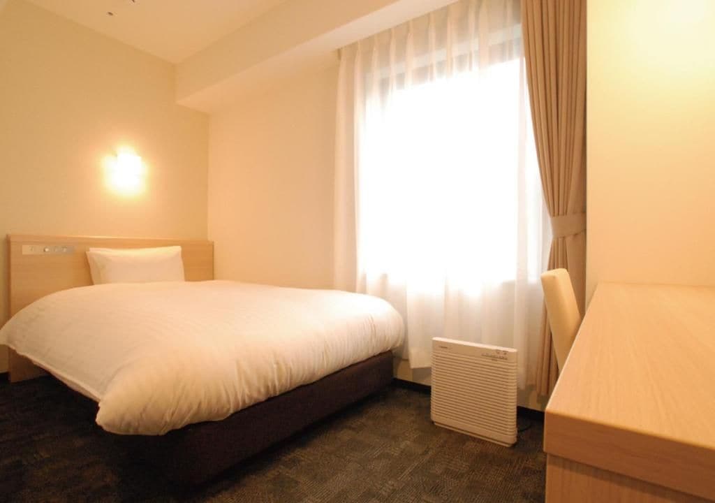 A hotel room with a bed, a desk, a radiator, and a window with curtains.