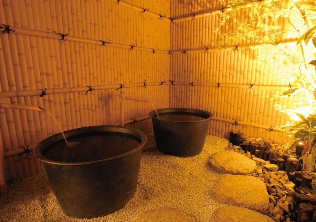 A room with a bamboo wall, two large metal buckets, and a small garden area with rocks and plants.