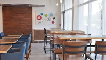A modern dining area with wooden tables and chairs, a wall-mounted art display, and large windows allowing natural light.