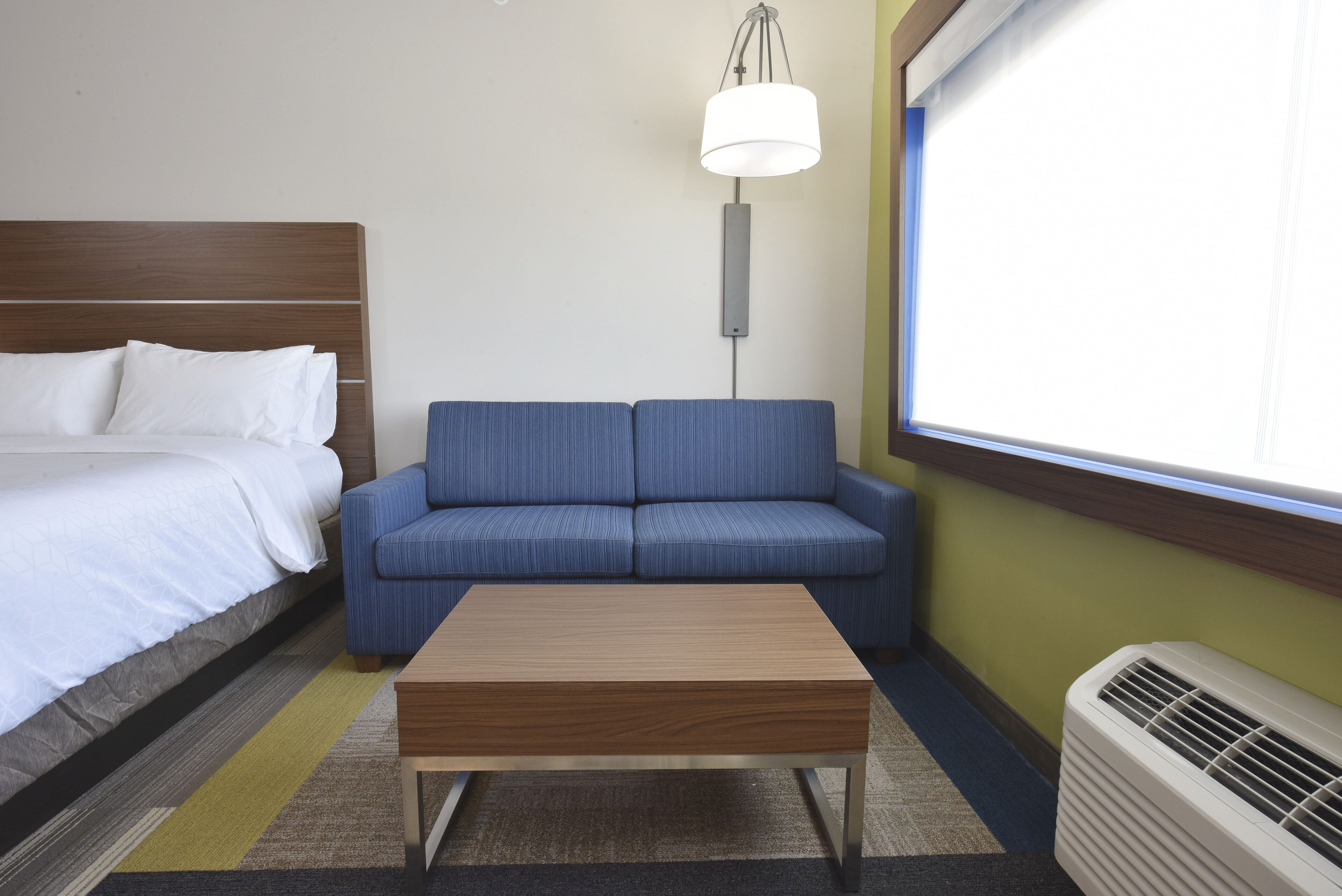 A hotel room with a bed, a blue sofa, a wooden coffee table, a floor lamp, and a window with blinds.