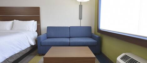 A hotel room with a bed, a blue sofa, a wooden coffee table, a floor lamp, and a window with blinds.