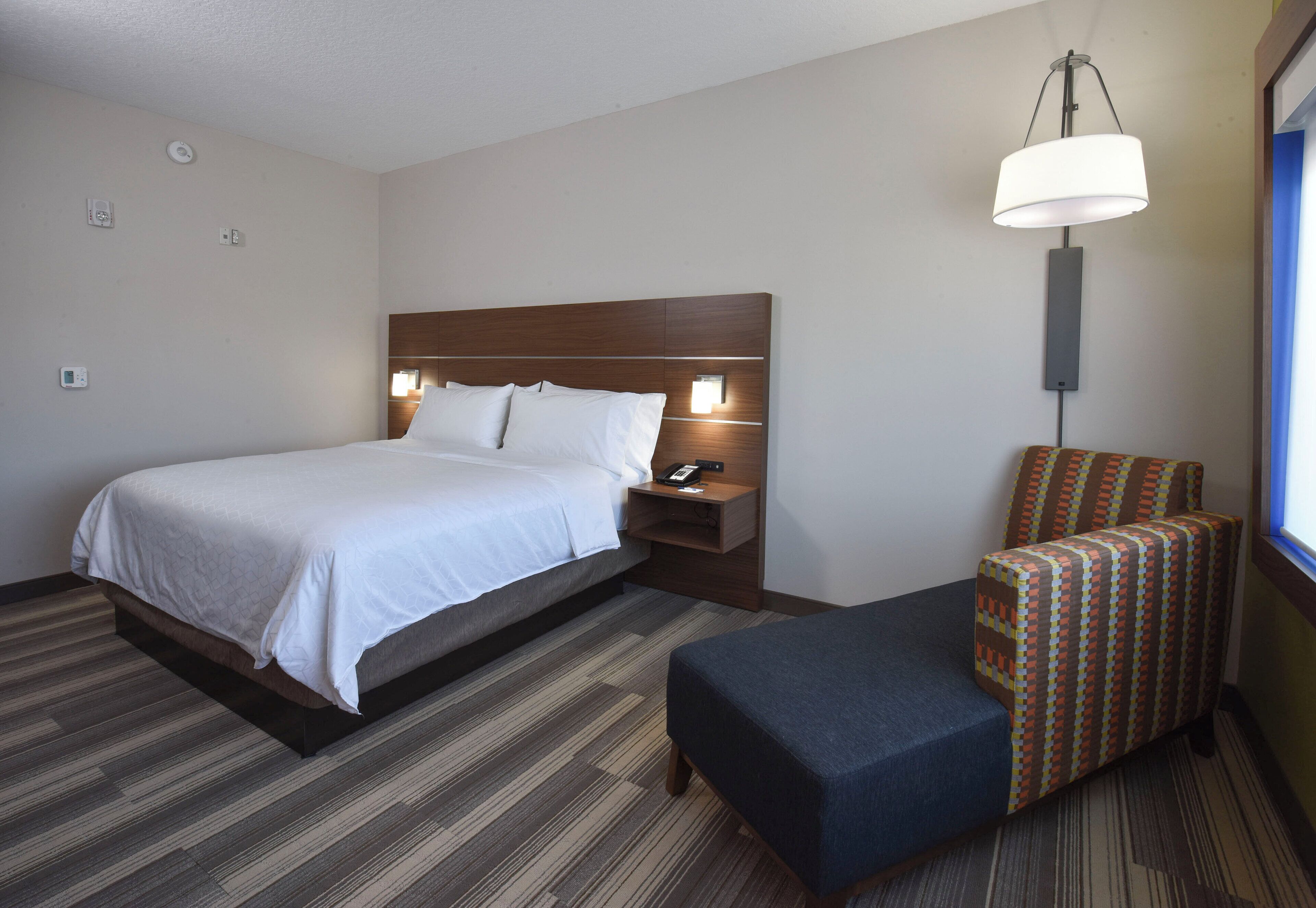 A hotel room with a bed, bedside table, lamp, and a patterned armchair.