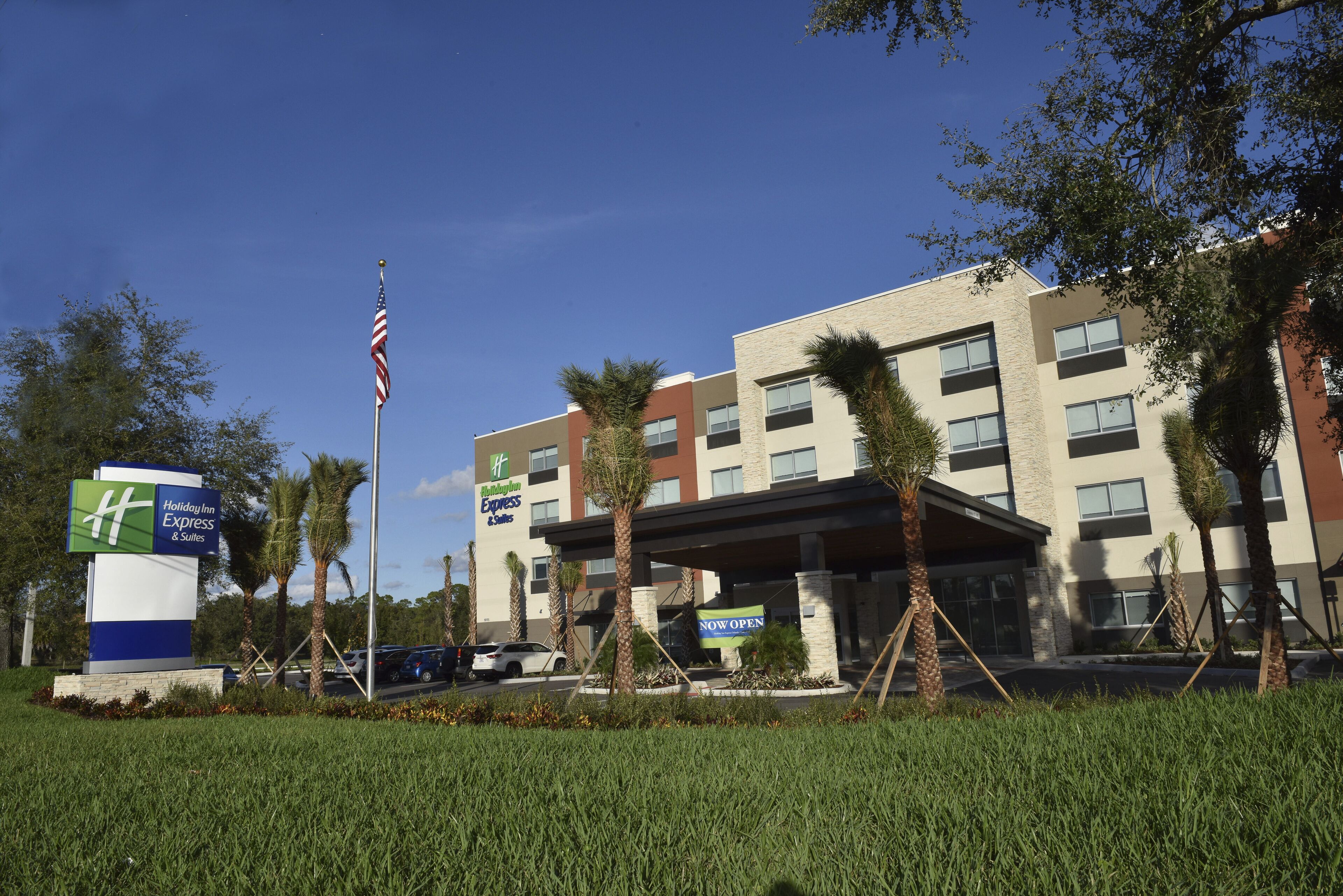 A Holiday Inn Express & Suites building with a 'NOW OPEN' sign, surrounded by palm trees and a clear blue sky.