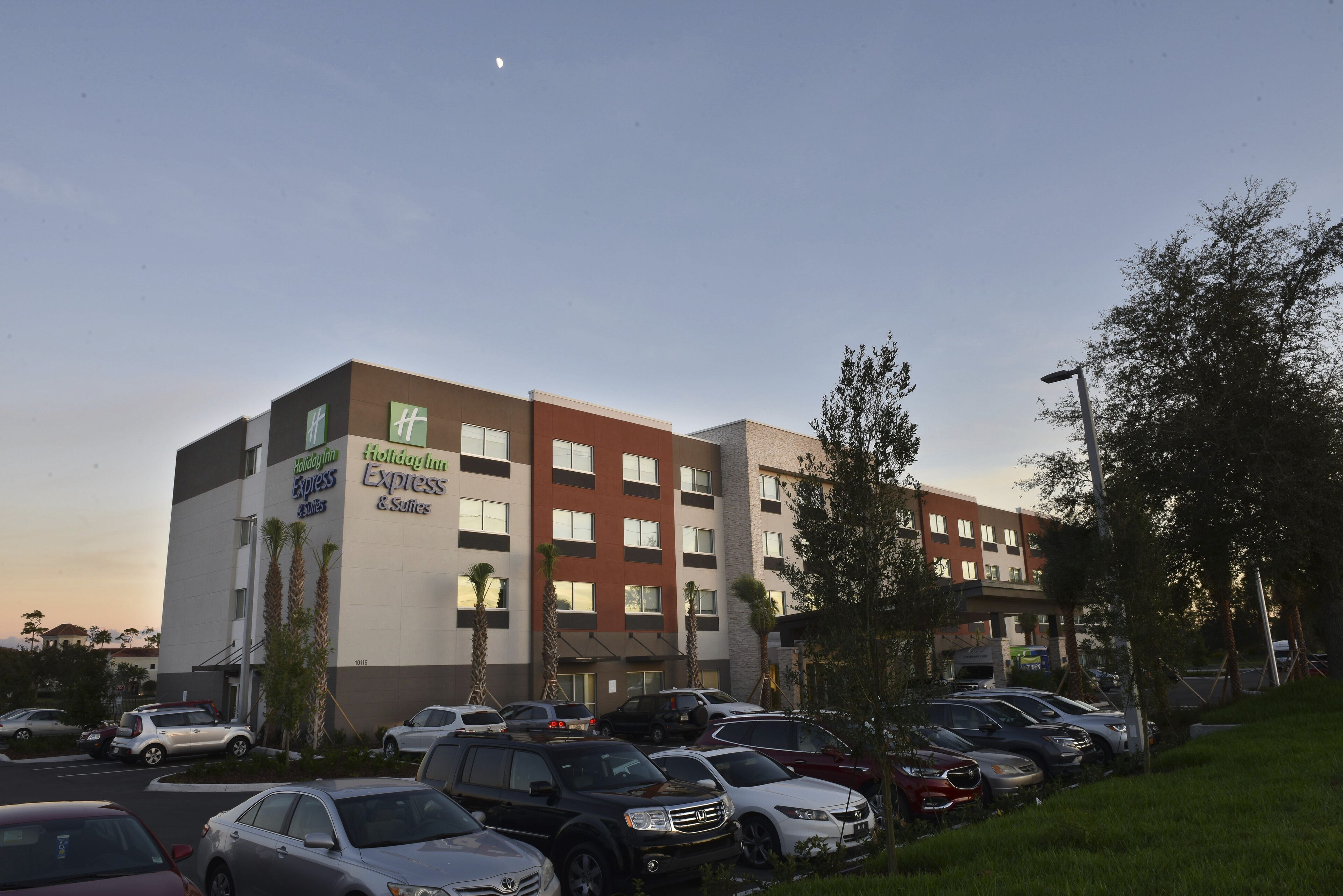 Un hotel Holiday Inn Express & Suites con un estacionamiento lleno de autos, rodeado de árboles y un cielo despejado.
