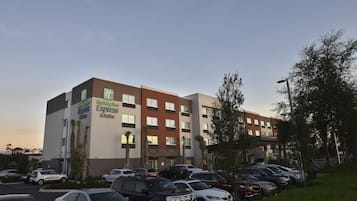 Un hotel Holiday Inn Express & Suites con un estacionamiento lleno de autos, rodeado de árboles y un cielo despejado.