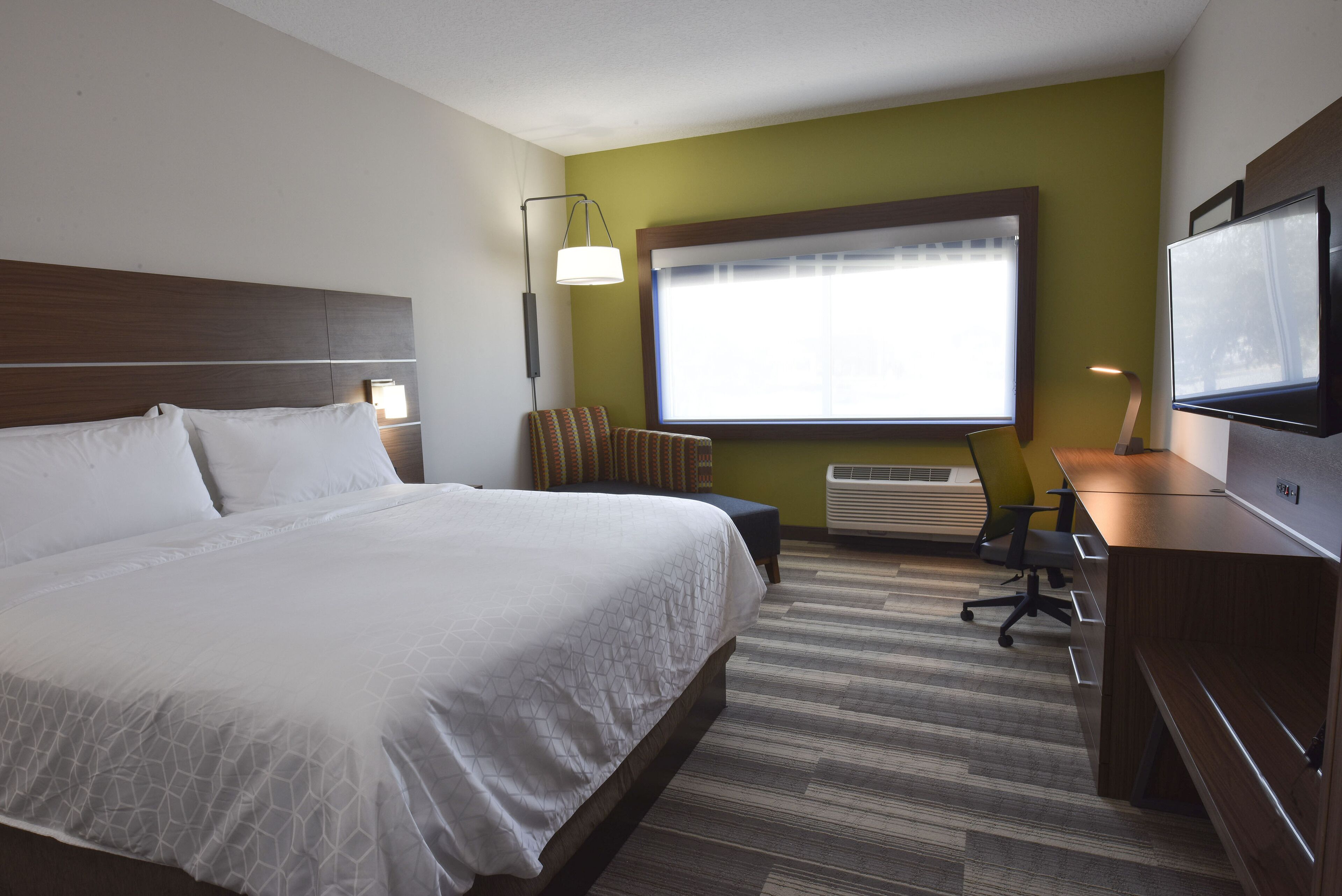 A hotel room with a large bed, a desk, a chair, a TV, and a window with blinds.