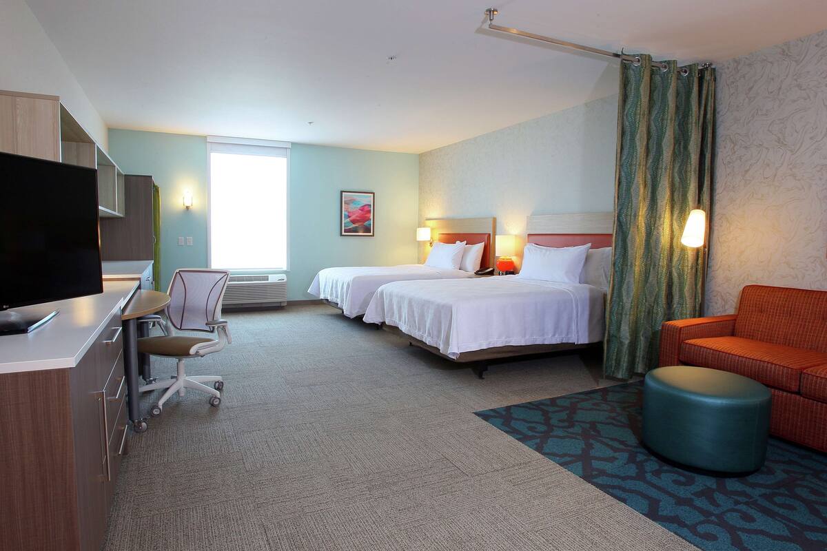 A hotel room with two beds, a desk with a TV, a chair, an ottoman, and a patterned rug.