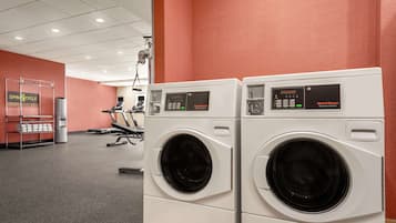 A laundry facility with two front-loading washing machines, a rack of folded towels, and a treadmill in the background.