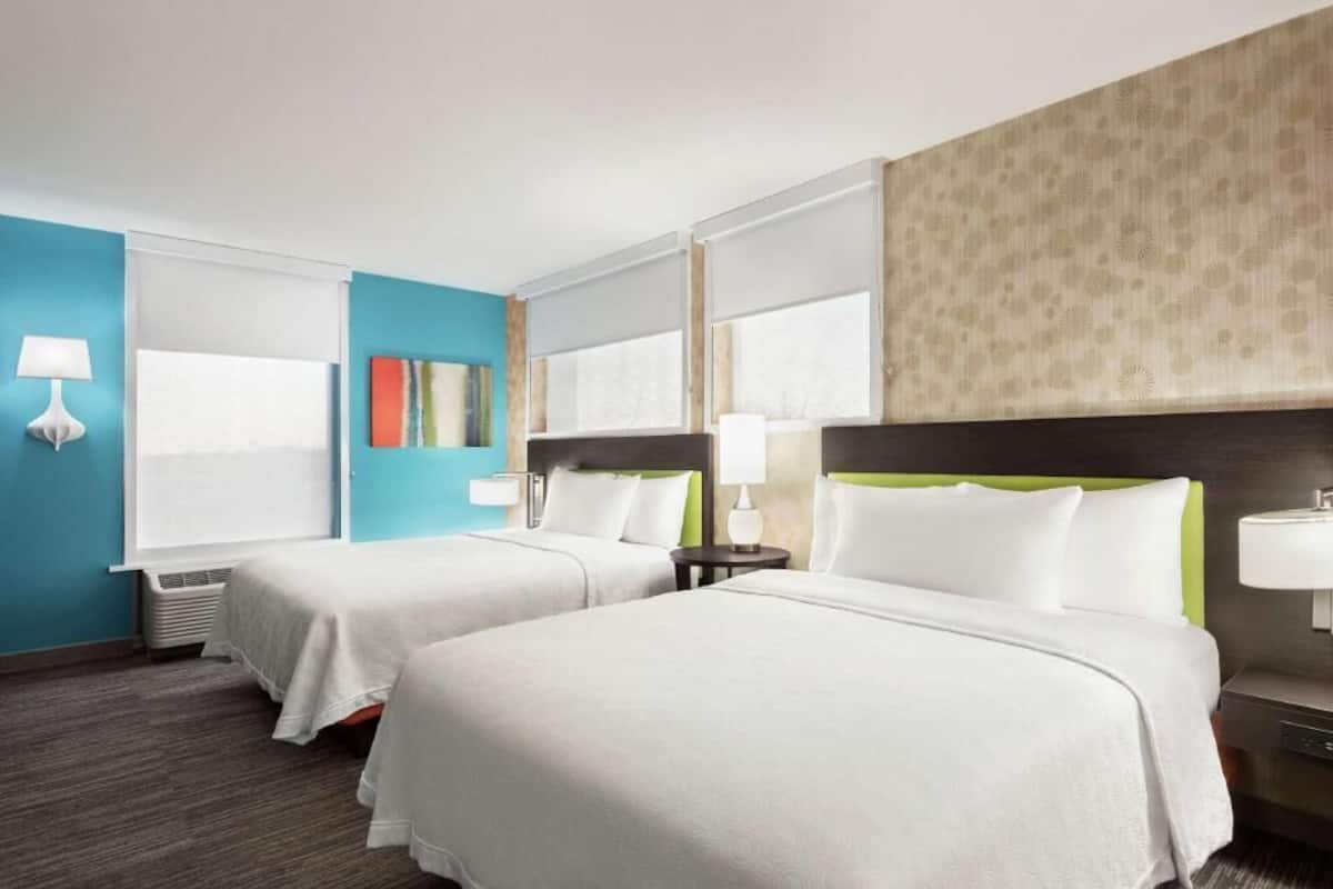 Ein Hotelzimmer mit zwei Betten, einem großen Fenster, einer Wand-Klimaanlage und einem Nachttisch mit Lampe.