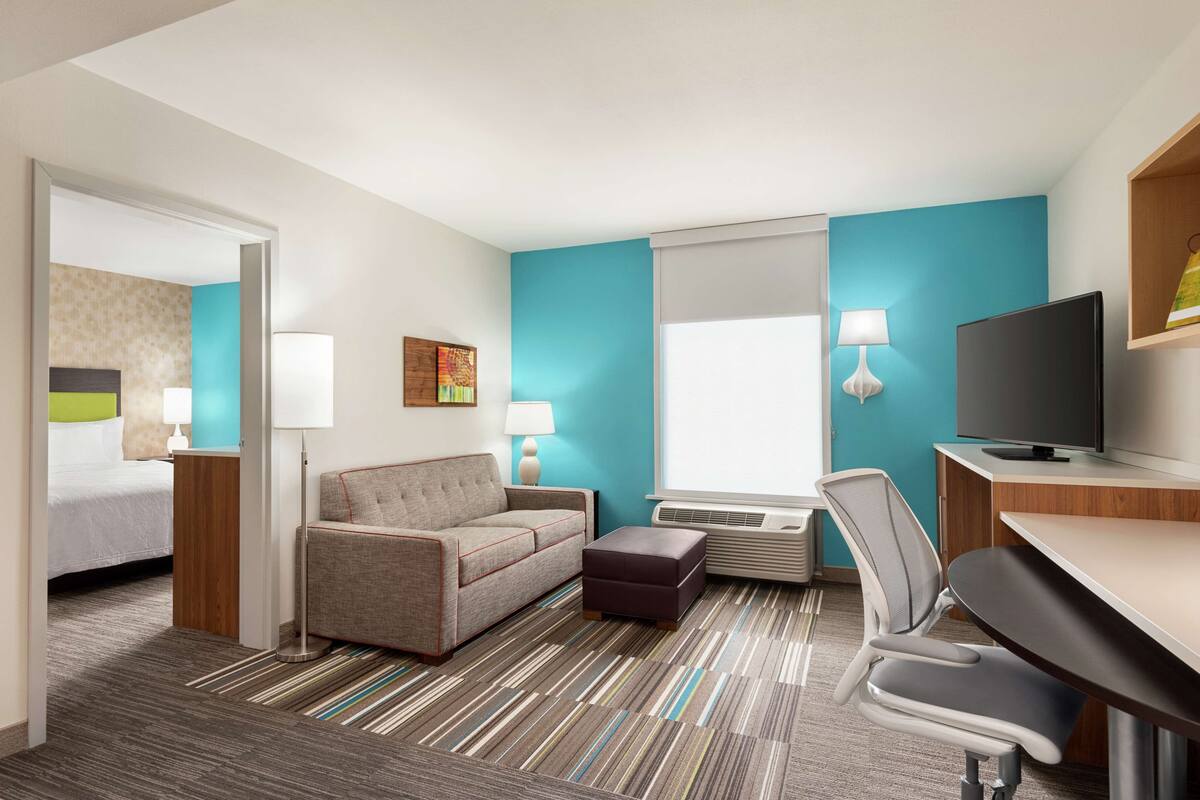 Ein Hotelzimmer mit Bett, einer Couch, einem Schreibtisch, einem Fernseher und einem Fenster mit Jalousien.