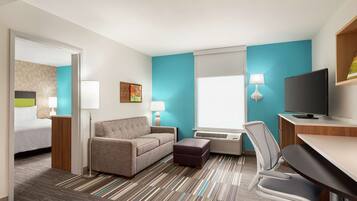 A hotel room with a bed, a sofa, a desk, a TV, and a window with blinds.