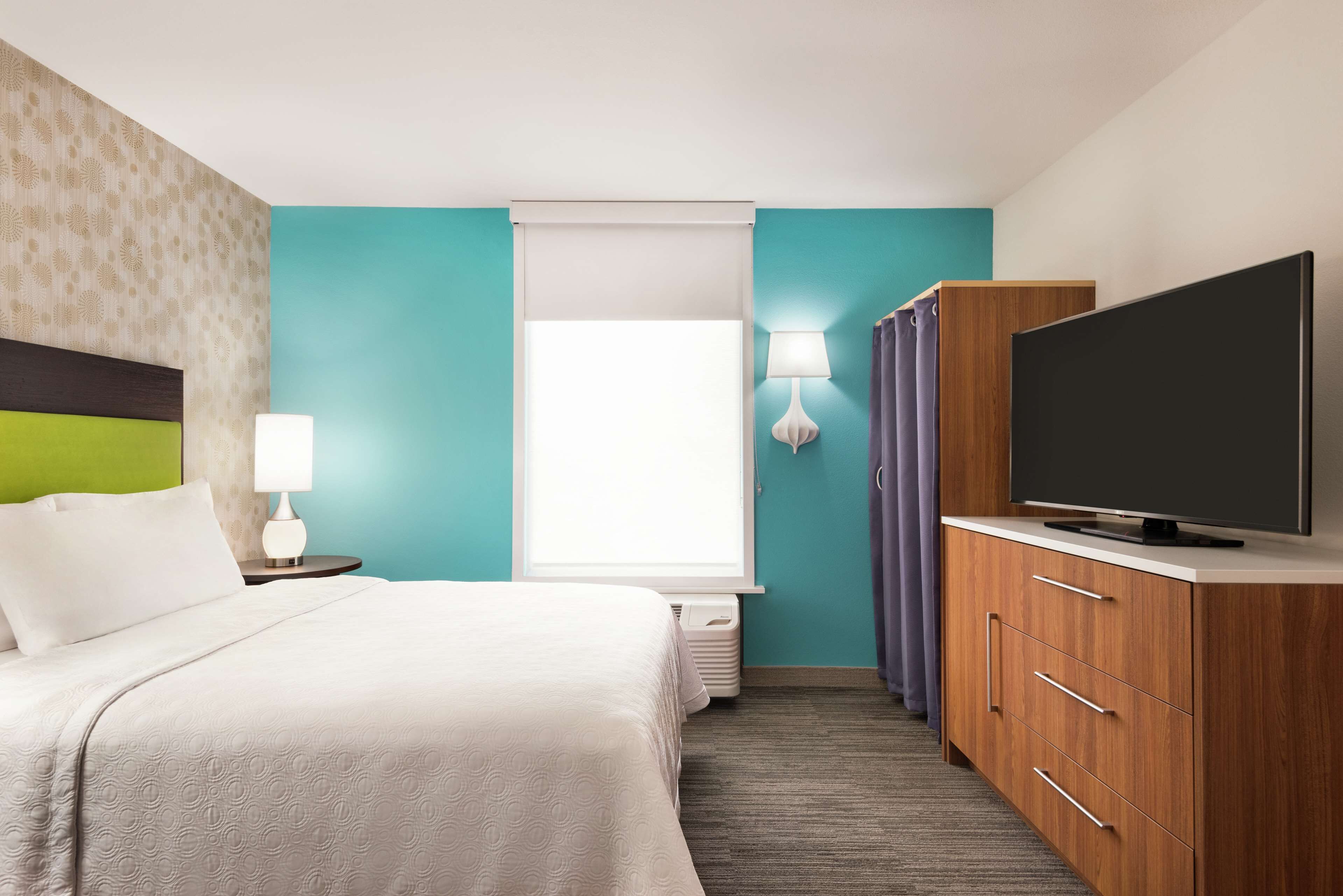 A hotel room with a bed, a TV, a nightstand, a lamp, a window with curtains, and a wall with a patterned design.