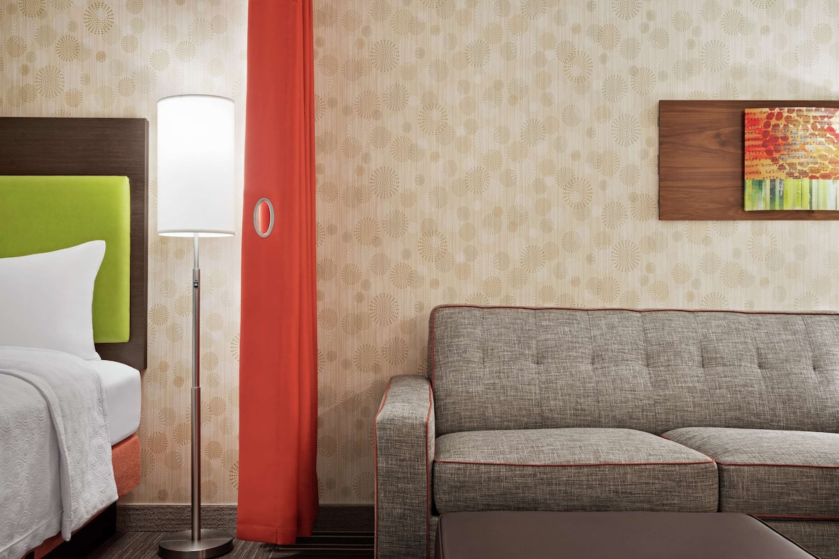 Ein Hotelzimmer mit einem Bett, einem grauen Sofa, einem roten Vorhang, einer Stehlampe und einer Wand mit strukturierter Tapete und einem gerahmten Kunstwerk.
