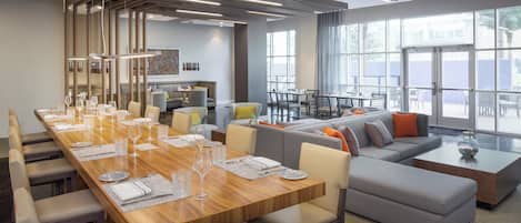 A modern dining area with a long wooden table set for a meal, a seating area with sofas and chairs, and large windows providing natural light.