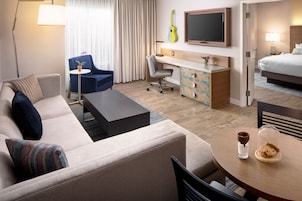 Habitación de hotel con sofá, escritorio, silla, cama, televisión y mesa de comedor con vaso y galleta.