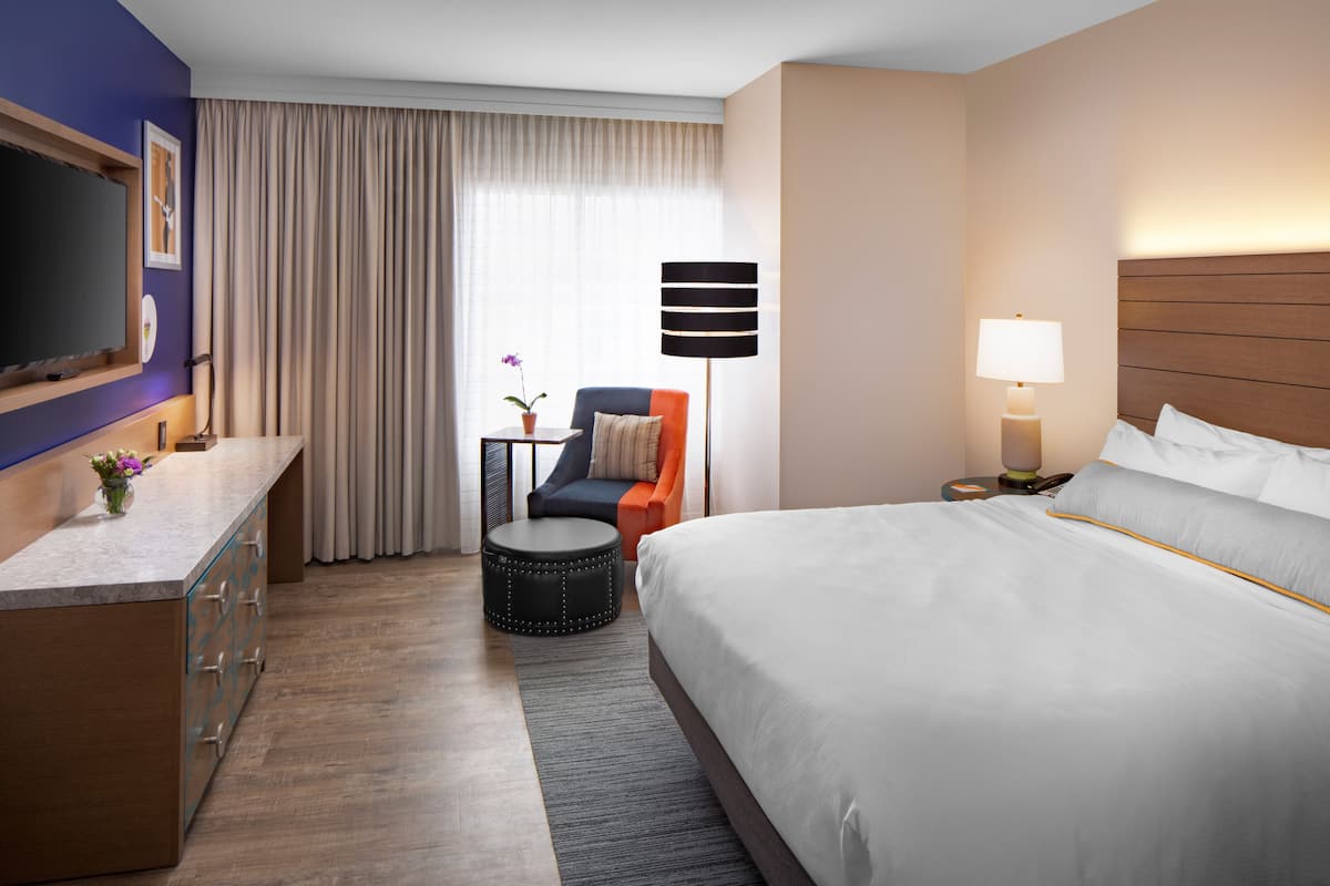 Une chambre d’hôtel avec un grand lit, un bureau doté d’un téléviseur à écran plat, une chaise, une petite table ronde avec une lampe et une fenêtre avec des rideaux.