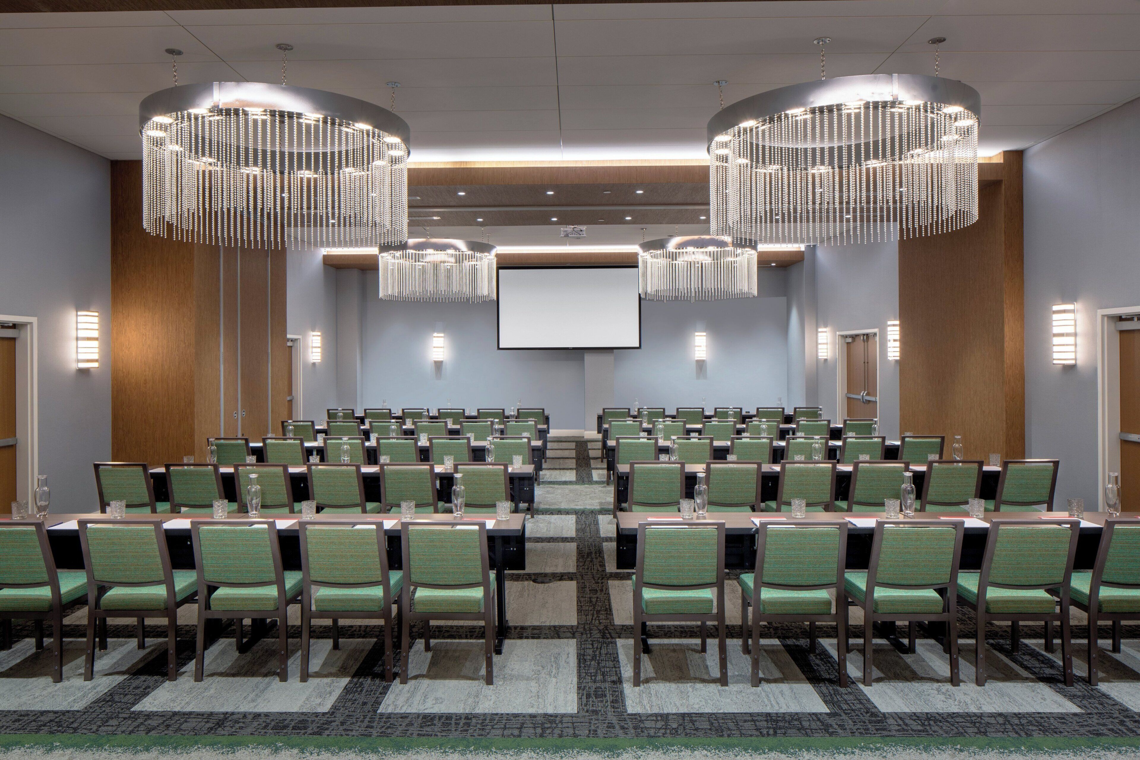 A conference room with rows of green chairs, a large screen, and two chandeliers.