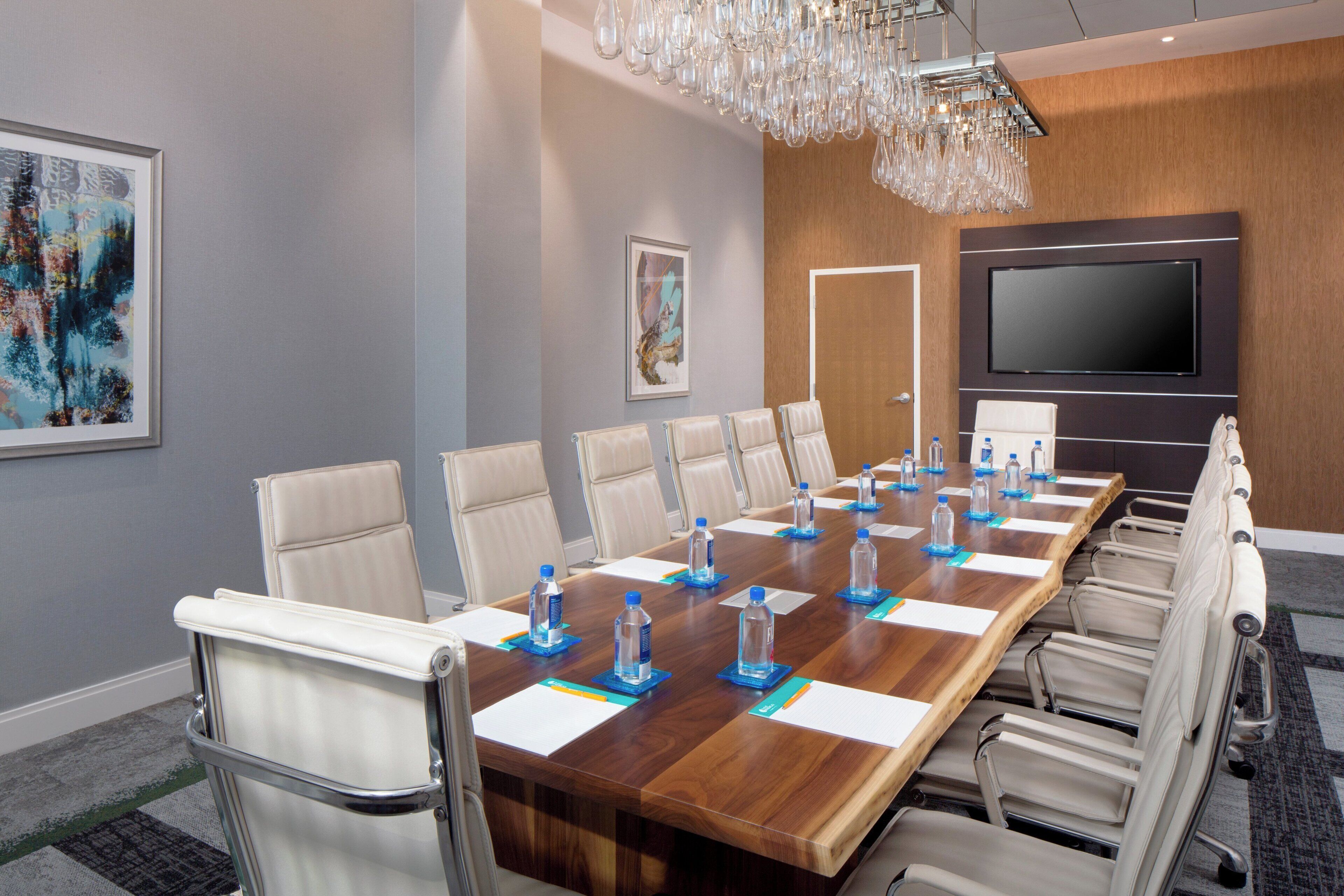 A conference room with a long wooden table, beige chairs, water bottles, and a large flat-screen TV on the wall.