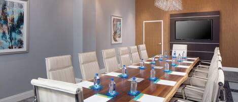 A conference room with a long wooden table, beige chairs, water bottles, and a large flat-screen TV on the wall.