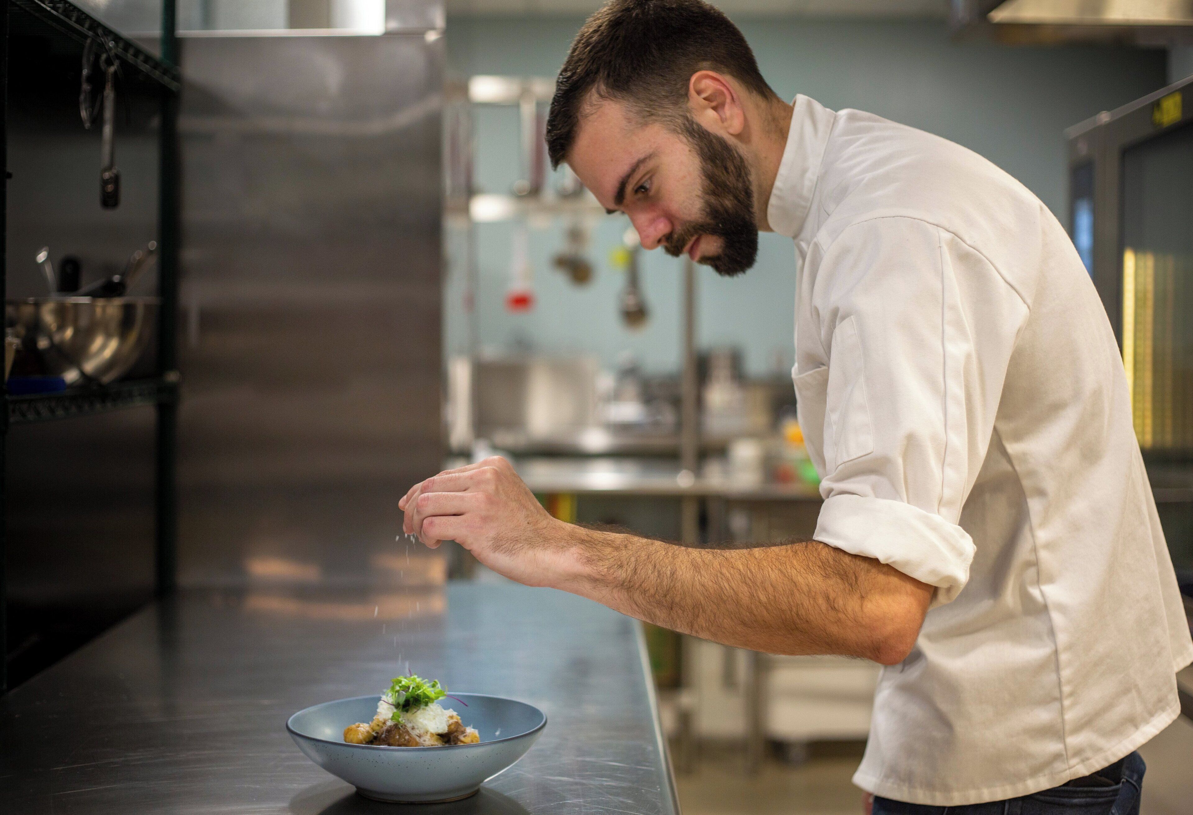 A chef in a white shirt preparing food in a kitchen with stainless steel appliances and a metal bowl on the counter.