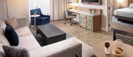 Habitación de hotel con sofá, escritorio, silla, cama, televisor y mesa de comedor con vaso y galleta.