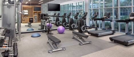 A modern gym with various exercise equipment, large windows, and a flat-screen TV.