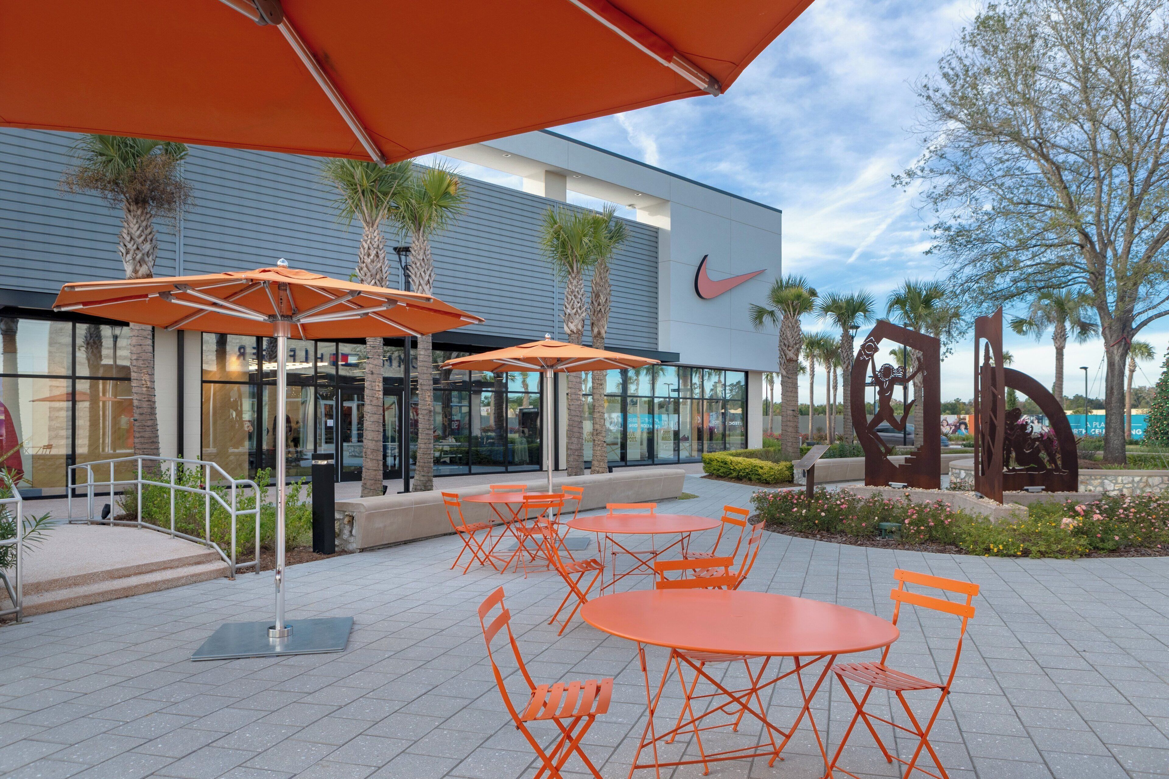Un espace extérieur aménagé avec des tables et des chaises orange, un bâtiment moderne orné du logo Nike et des palmiers.