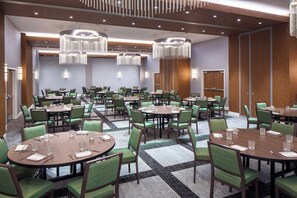 En restaurant i et hotel med flere runde borde, grønne polstrede stole og moderne lysekroner.