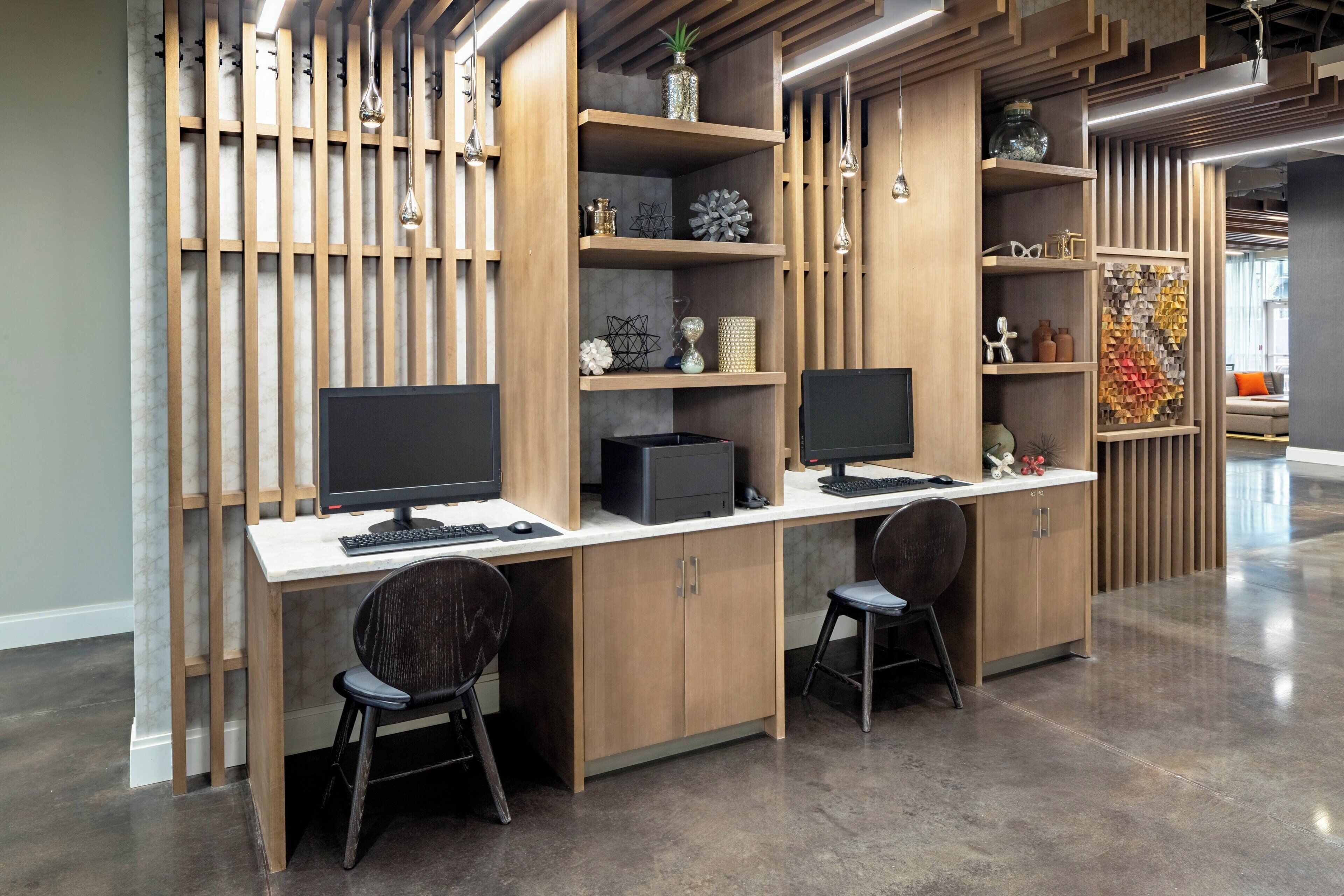 A modern office space with two desks, each with a computer monitor, keyboard, and mouse. There are two chairs, a decorative wall with shelves, and a patterned floor.