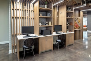 A modern office space with two desks, each with a computer monitor, keyboard, and mouse. There are two chairs, a decorative wall with shelves, and a patterned floor. - Hotel Indigo Gainesville-Celebration Pointe by IHG (Gainesville)