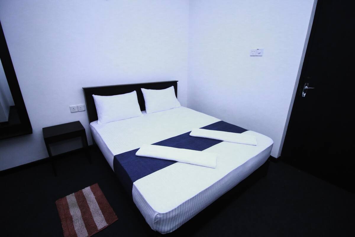 Una cama bien tendida con ropa de cama blanca y azul oscuro, una mesita de noche pequeña y una alfombra a rayas sobre un suelo oscuro.