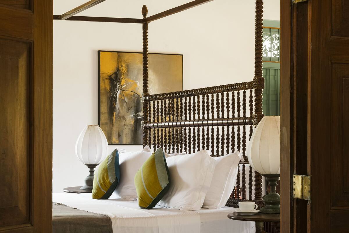 A four-poster bed with white linens and green and yellow pillows, a wooden floor, and a painting on the wall.