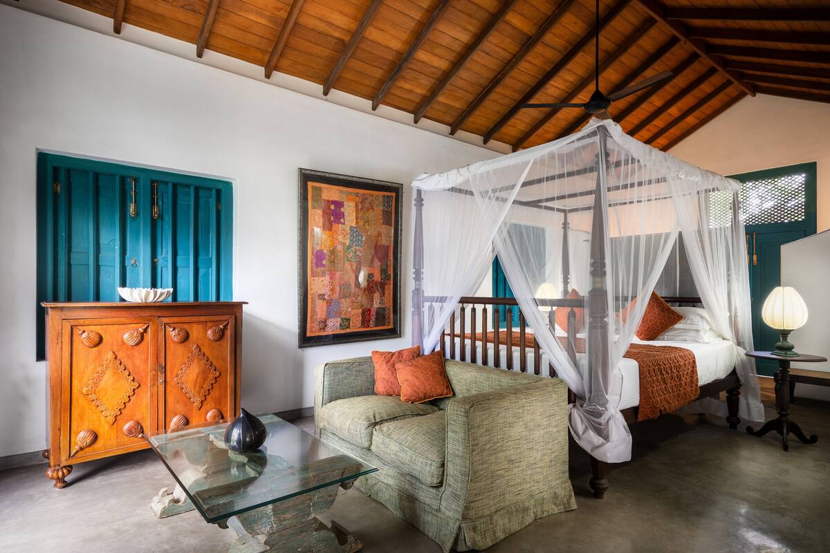A room with a wooden ceiling, a bed with a canopy, a sofa, a glass table, a cabinet, and a painting on the wall.