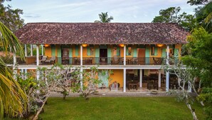 A two-story building with a tiled roof, green shutters, and a balcony with wooden railings. - The Last House (Morakatiyara)