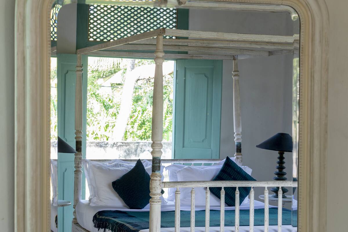 A four-poster bed with white and blue bedding, a green door, and a white wall with a decorative mirror.