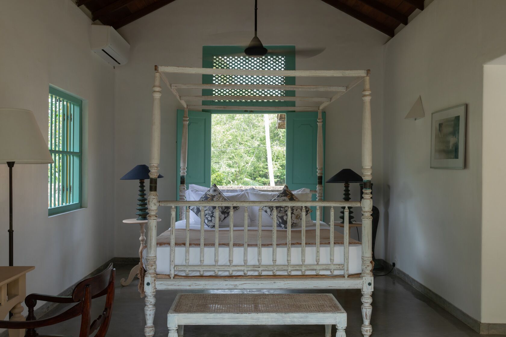 A four-poster bed with a bench in front, a chair, a lamp, and a window with shutters.