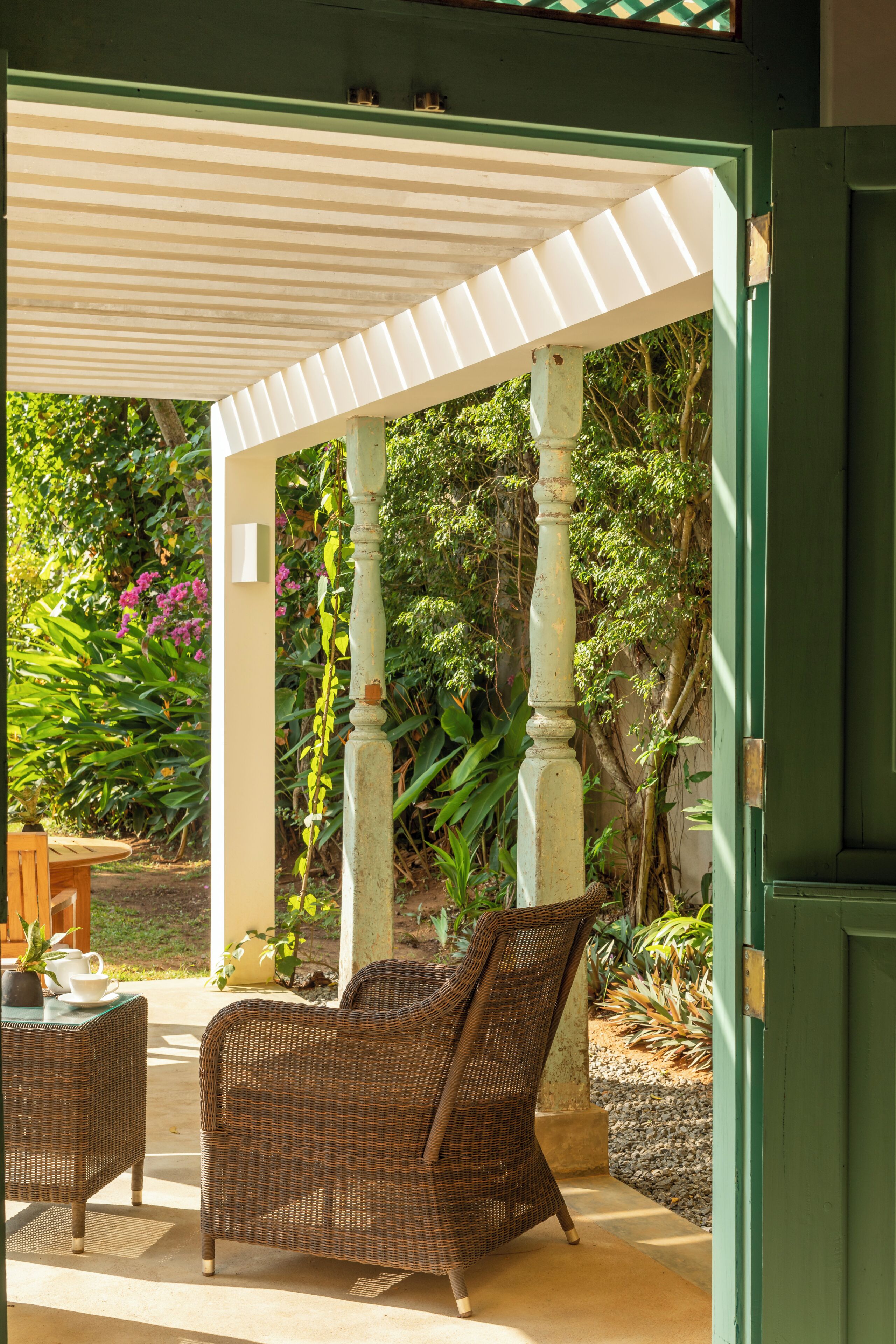 An outdoor seating area with wicker furniture under a white pergola, surrounded by greenery and a stone wall.