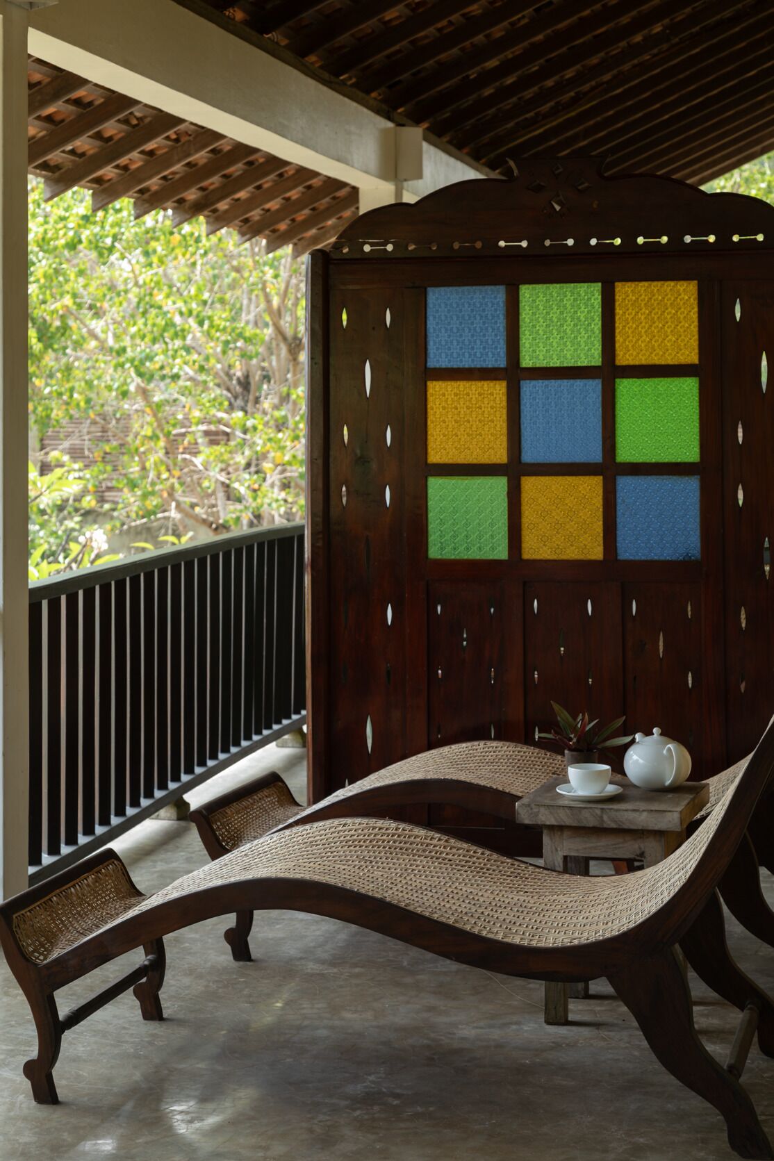 A wooden bench with woven seats and a table with a teapot and cup, set against a wooden partition with a grid of colored panels.