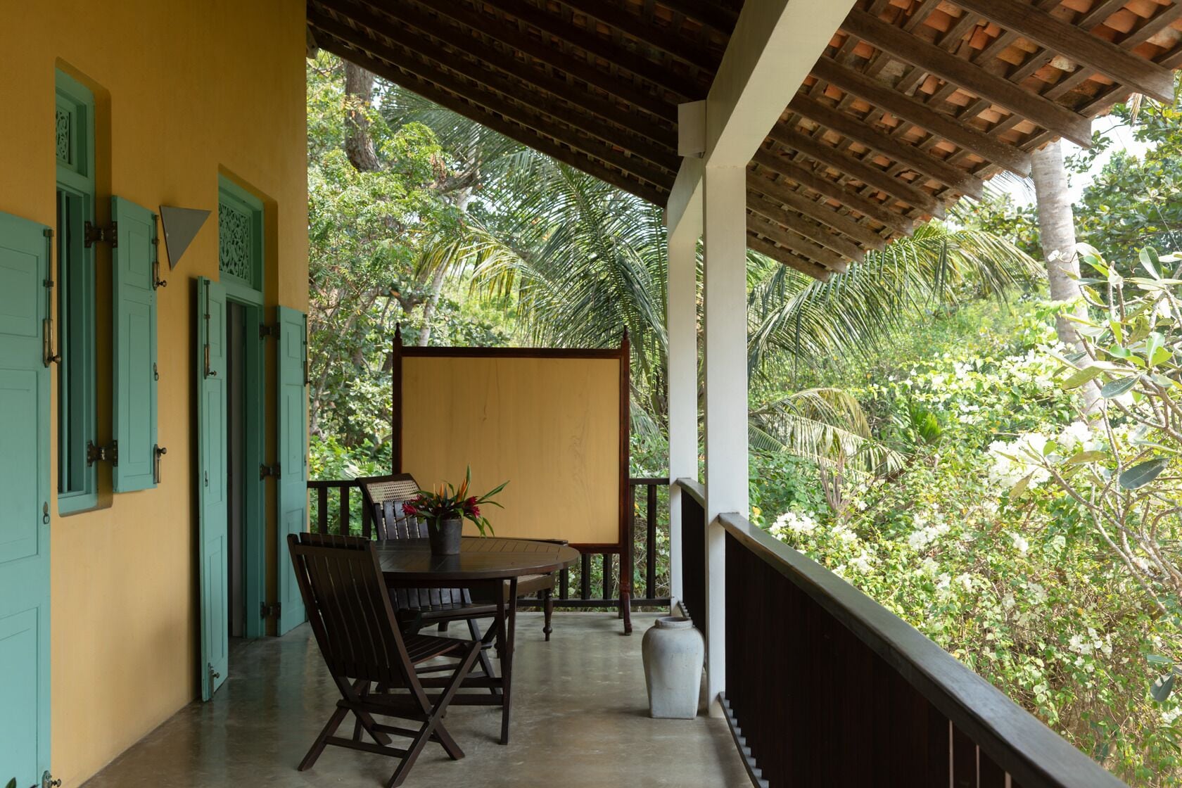 A covered outdoor seating area with a table and chairs, surrounded by lush greenery.
