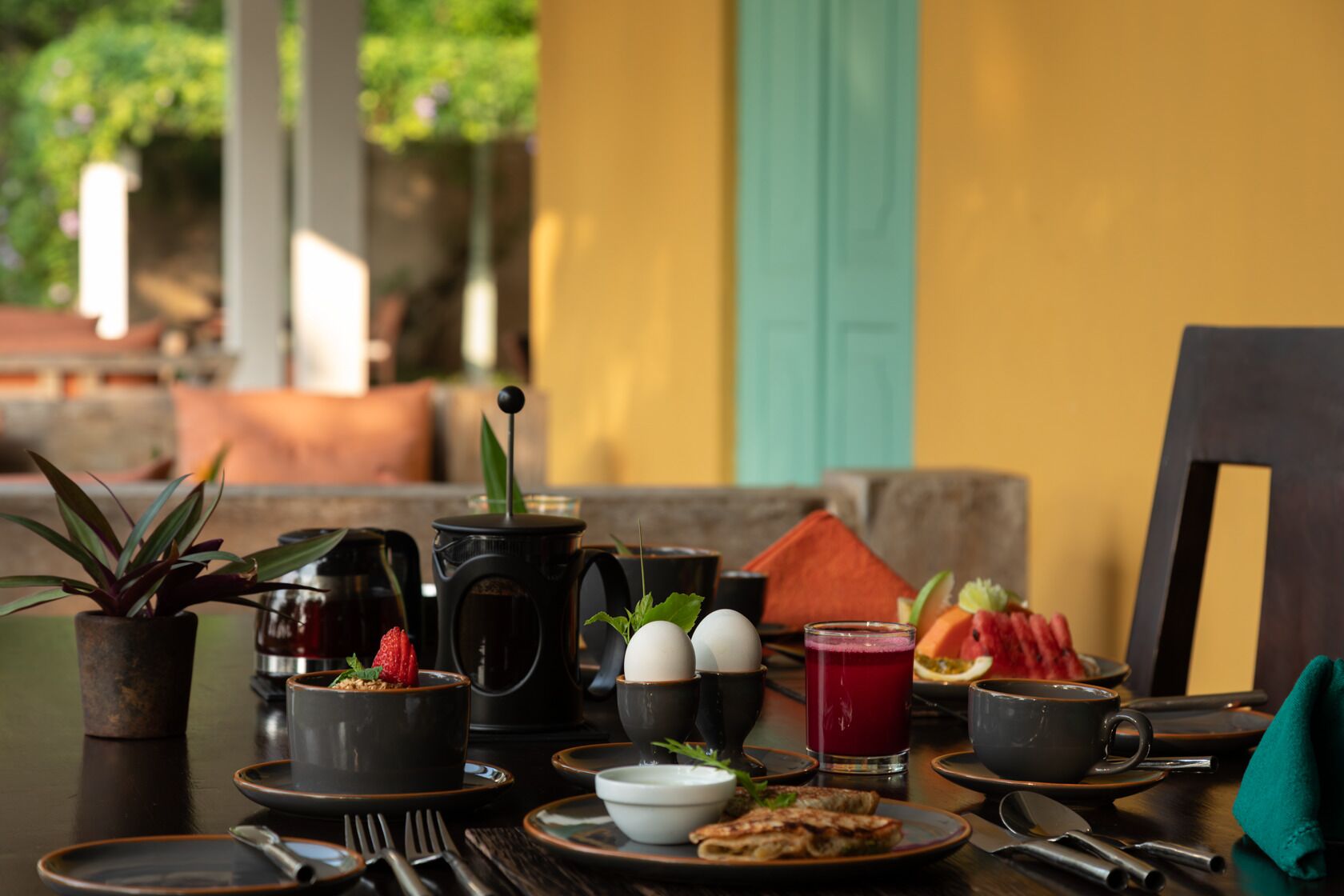A table set with breakfast items including eggs, toast, fruit, and beverages, with a view of greenery outside.