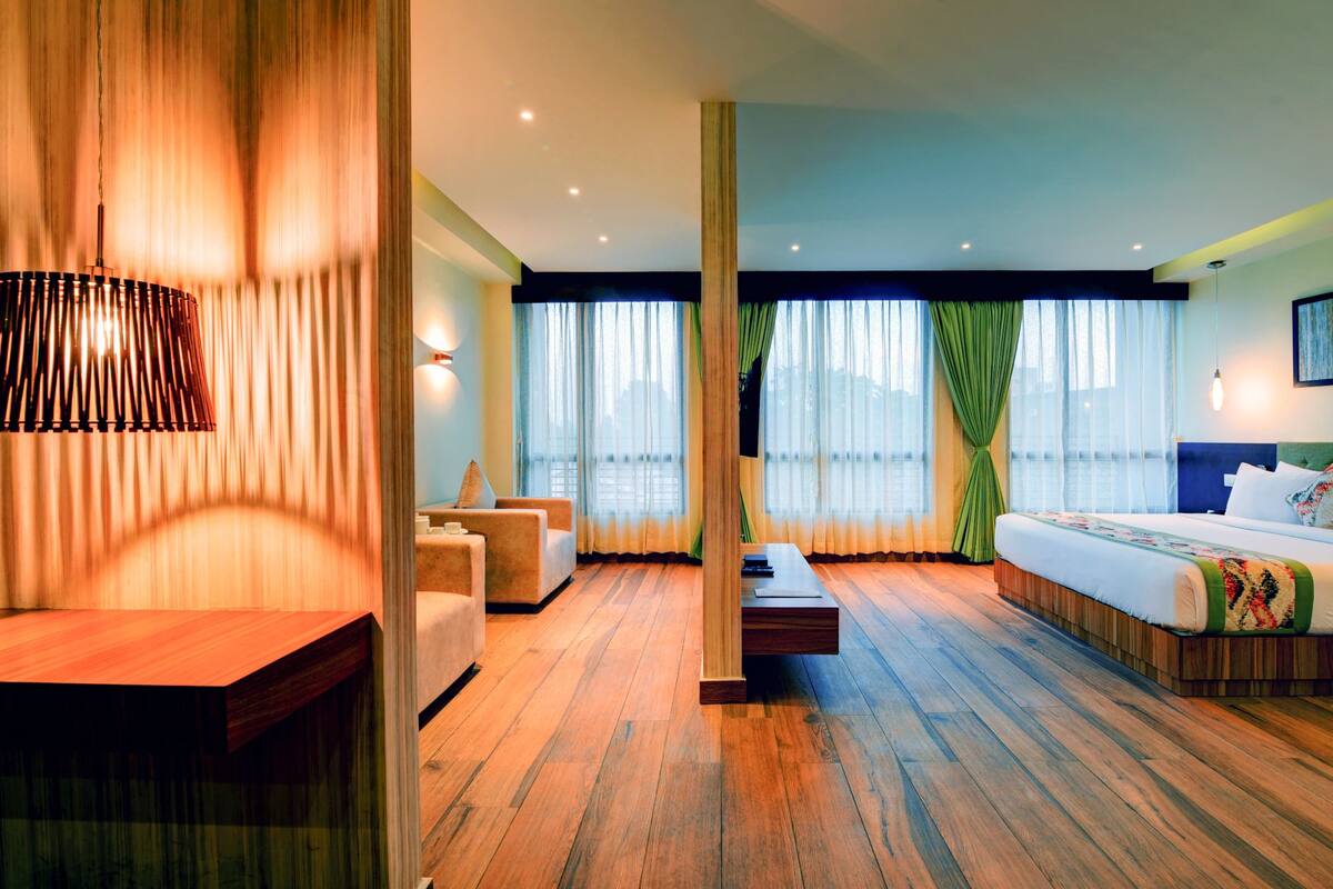 Habitación de hotel con suelo de madera, cama con cubrecama verde y blanco, escritorio con lámpara y vistas de la ciudad a través de amplias ventanas.
