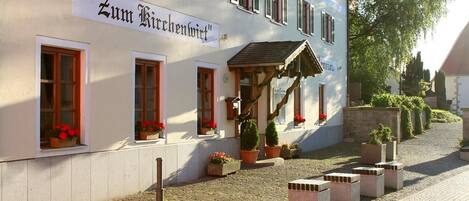 A building with a sign reading "Zum Kirchenviertel". It has multiple windows with flower boxes, a small porch with a roof, and potted plants around the entrance.