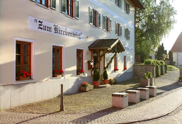 A building with a sign reading "Zum Kirchenviertel". It has multiple windows with flower boxes, a small porch with a roof, and potted plants around the entrance.