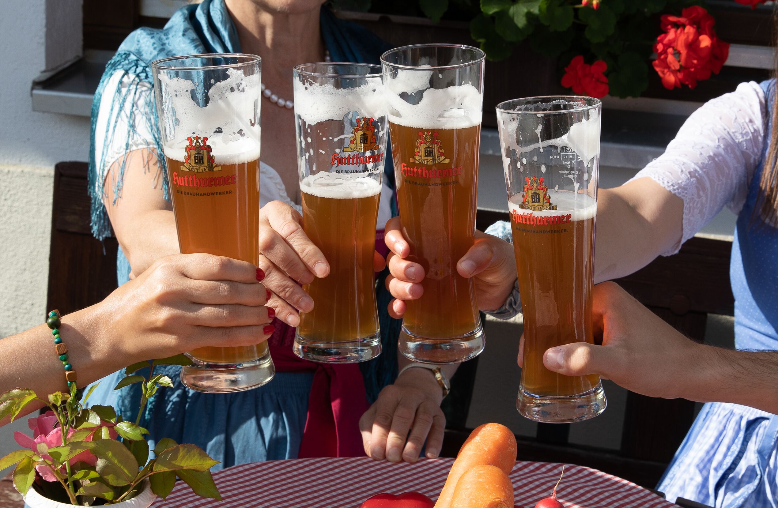 Four people toasting with glasses of beer at a table with a red and white checkered tablecloth, surrounded by flowers and vegetables.