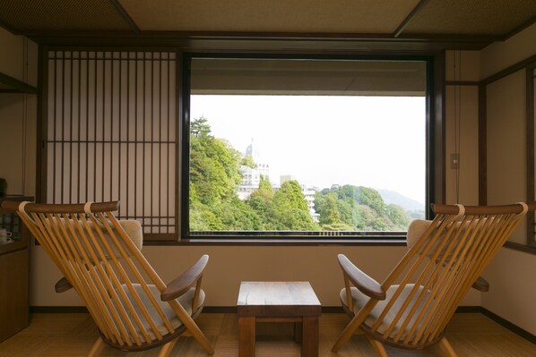 A room with two wooden chairs and a small table, a window with a view of trees and buildings, and traditional sliding doors.
