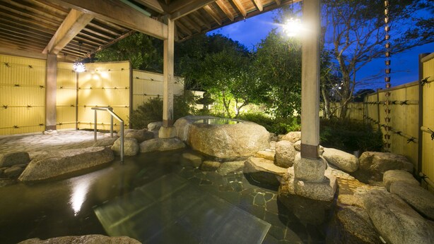 A traditional Japanese onsen with a wooden roof, stone bath, and a small wooden staircase leading into the water.