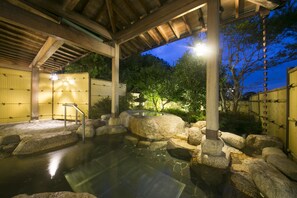 Un onsen japonais traditionnel, avec un toit en bois, un bain en pierre et un petit escalier en bois menant à l’eau.