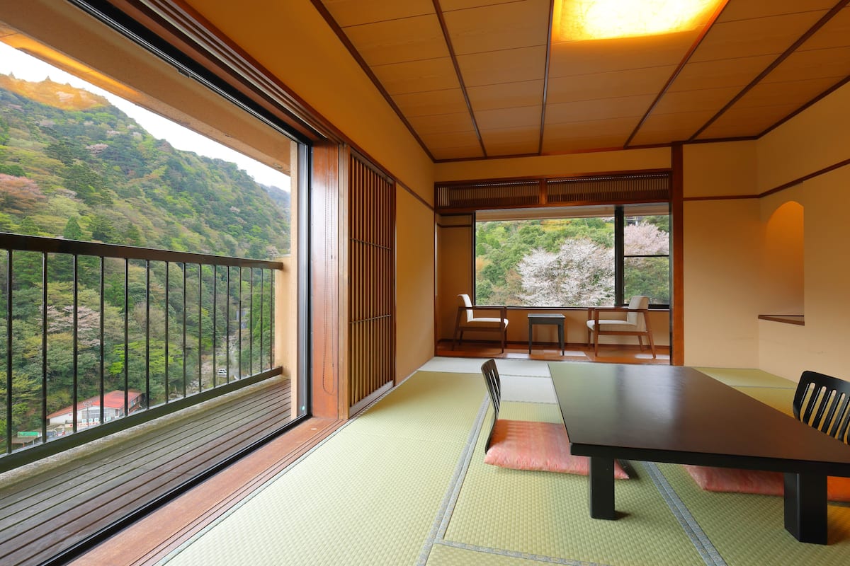 Une pièce de style japonais traditionnel, avec un sol recouvert de tatamis, une table basse et une vue sur un paysage montagneux grâce à de grandes fenêtres.