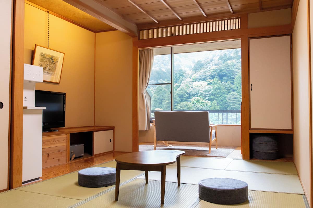 Une pièce de style japonais traditionnel, avec un sol recouvert de tatamis, un téléviseur, une porte coulissante donnant sur un balcon et une vue sur une zone boisée.